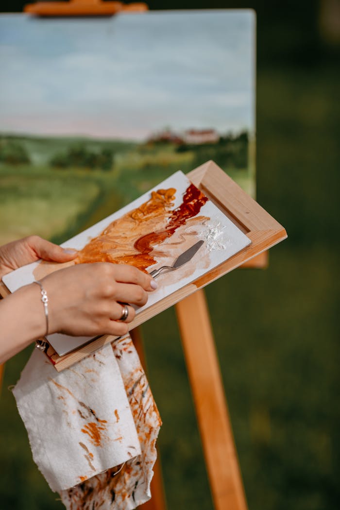 Artist holds palette while painting landscape outdoors, blending vibrant colors.